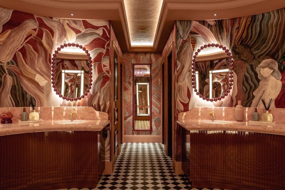 A pink bathroom with led backlit mirrors, bold botanical pink and green wallpaper with pink marble vanities