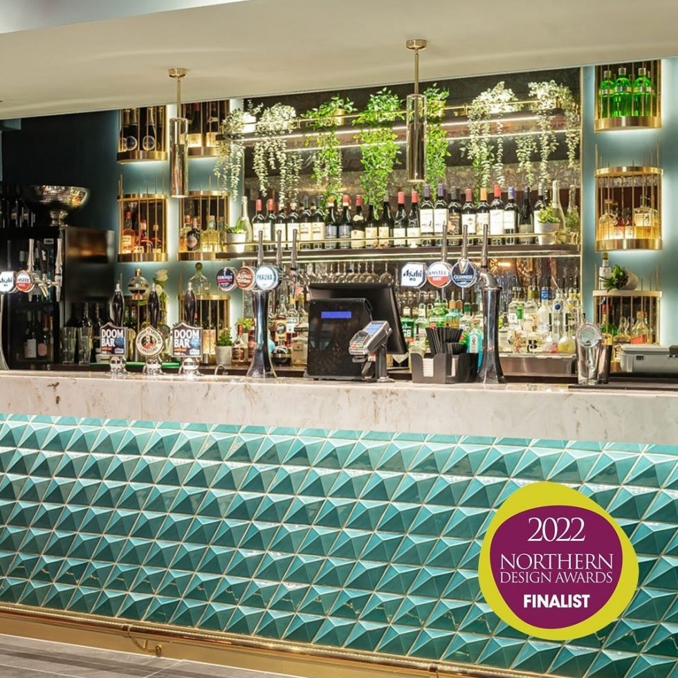 Green and gold colour themed bar with green shaped tiles to front of bar. White marble with gold vein bar top - plants and greenery to the rear of the bar