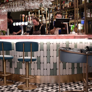 a man and woman behind the bar, the bar has a pink marble bar top and blue and white tiles running down the bar walls with blue high chairs surrounding the bar