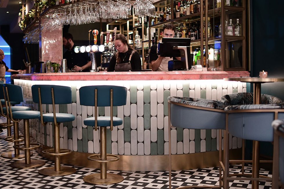 a man and woman behind the bar, the bar has a pink marble bar top and blue and white tiles running down the bar walls with blue high chairs surrounding the bar