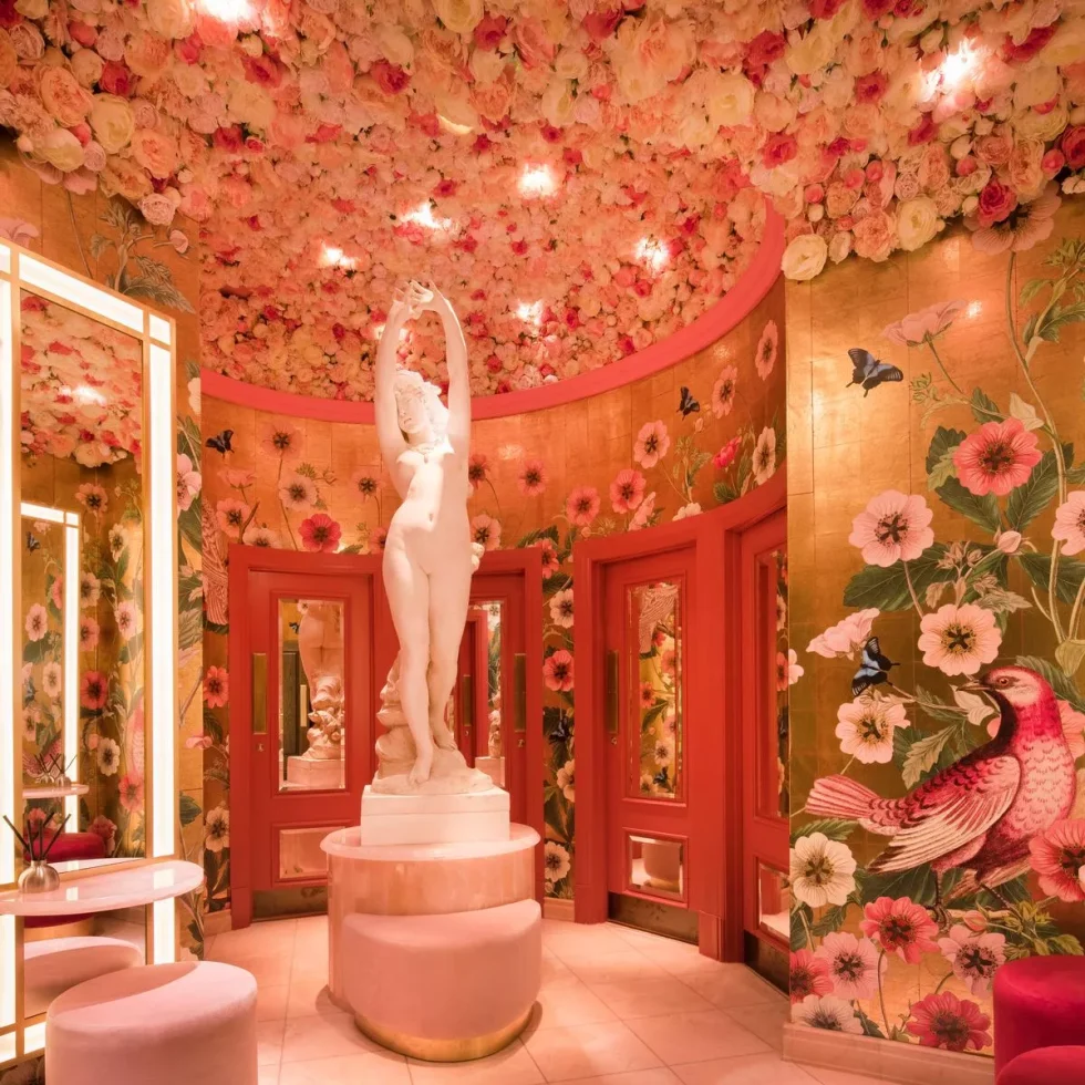 Bold pink and gold washrooms with large floral wallpaper and a statue of a woman in the washroom stood upon a pink marble stand.